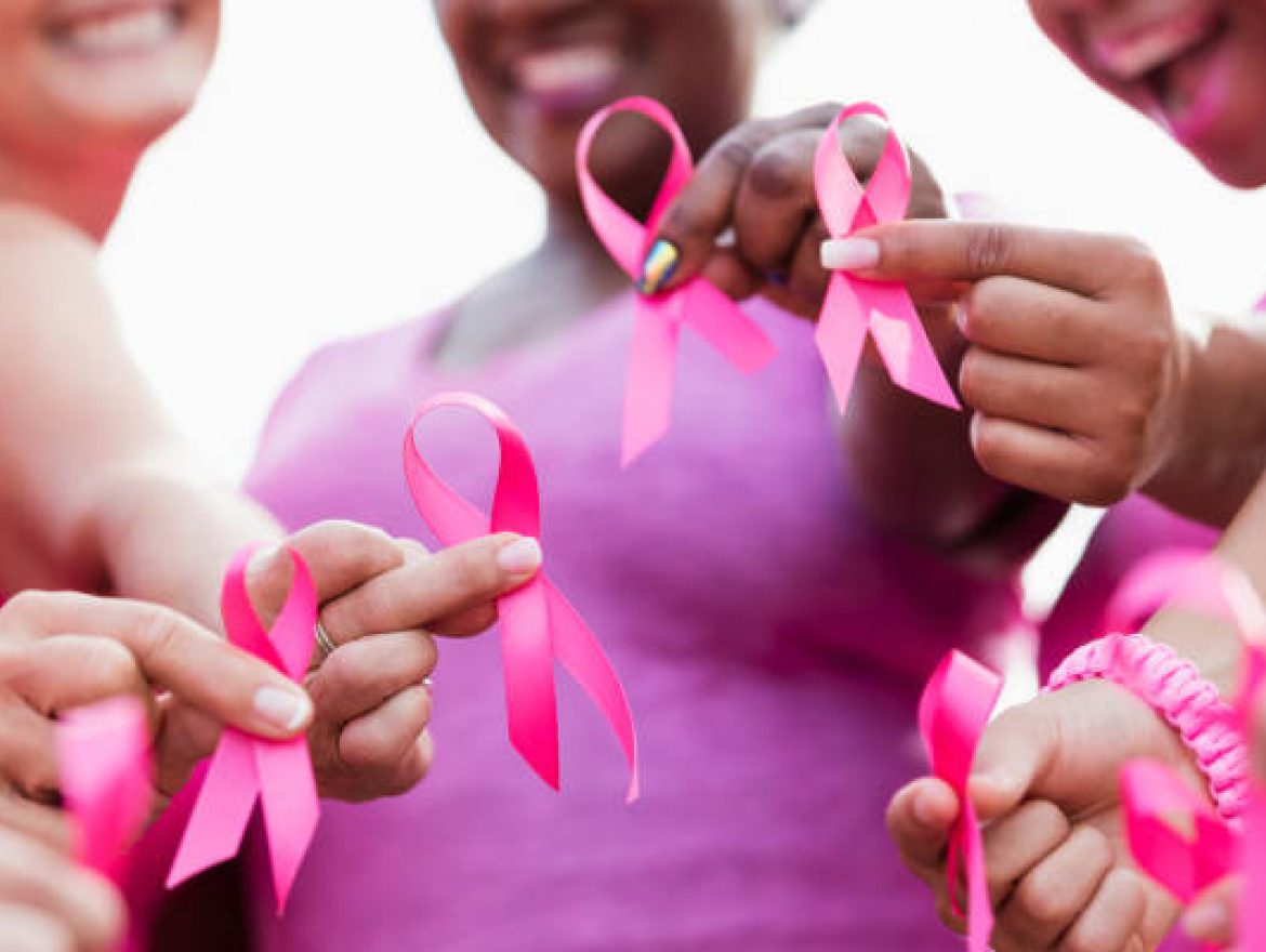 Cropped view of a multi-ethnic group of women of mixed ages standing together outdoors, wearing pink, at a breast cancer awareness rally, raising money to find a cure. They are each holding a breast cancer awareness ribbon. The focus is on their hands.